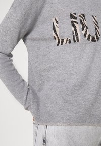 Szary sweter z długimi rękawami, z teksturowanym logo "LIU" w czarno-beżowy wzór zebry. Posiada prążkowany ściągacz u dołu oraz metalową metkę.