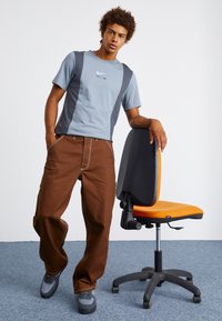Gråblå T-shirt med mörkgrå sidopaneler och vit "AIR"-logotyp, kombinerad med bruna byxor. Svarta och grå sneakers. Orange stol i bakgrunden.