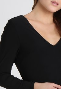 Femme portant un haut noir à manches longues avec un col en V, montrant une partie de son cou, de son épaule et de sa main sur un fond uni.