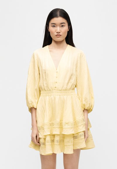 Νεαρή γυναίκα με μακριά σκουρόχρωμα μαλλιά, φορώντας ένα ανοιχτό κίτρινο φόρεμα με λαιμόκοψη σε σχήμα V, φουσκωτά μανίκια και φούστα με βολάν και δαντέλα, στέκεται μπροστά σε λευκό φόντο.