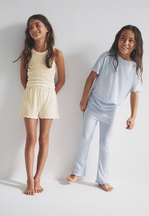 Deux filles souriantes aux longs cheveux sont debout pieds nus contre un mur blanc, vêtues de vêtements décontractés et amples de couleur jaune clair et bleu pâle.