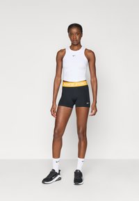 Fehér sport crop top fekete Nike logóval, fekete rövidnadrággal, amely sárga Nike Pro derékpánttal van párosítva. Fekete sportcipő és fehér zokni.