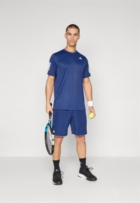 Tumed sinine spordiline tennisekomplekt lühikeste varrukatega, sobivad lühikesed püksid ja valged aktsendid. Mudel hoiab tennisereketit ja palli.