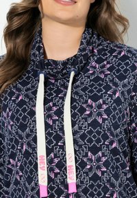 Marinblå huvtröja med ett geometriskt mönster i rosa och vitt. Den har en krage med vita dragsnören och rosa detaljer.