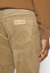 Smeđe kordone hlače s teksturiranom površinom, s kožnom oznakom Wrangler i karakterističnim šavovima na stražnjim džepovima.