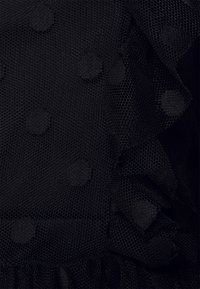 Tecidos de tule com bolinhas pretas, apresentando uma borda franzida. A textura é transparente, com bolinhas redondas salientes dispostas uniformemente.