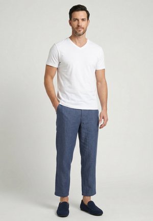 Mann steht mit Händen in den Taschen, trägt ein weißes kurzärmeliges V-Ausschnitt-Shirt, blaue gekürzte Hose und marineblaue Slip-On-Schuhe.