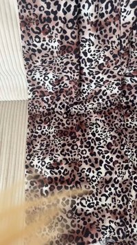 Leopardenmusterstoff mit glänzender Textur, der schwarze und braune Flecken auf einem cremefarbenen Hintergrund zeigt, über einer strukturierten Oberfläche drapiert.