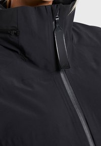 Gros plan sur une veste noire à col haut avec une fermeture éclair verticale et un tire-zip en cuir noir arborant un logo Adidas.