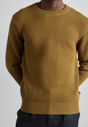 Homme portant un pull en tricot texturé jaune moutarde et un pantalon foncé, debout devant un fond clair uni.