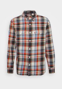 Chemise en flanelle à manches longues avec boutons, motif à carreaux rouge, bleu, noir et blanc, et deux poches poitrine avec boutons.