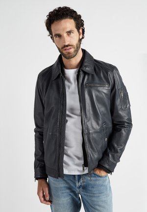 Man met krullend haar en baard, gekleed in een opengeknoopte zwart leren jas over een wit T-shirt en blauwe spijkerbroek, staand tegen een witte achtergrond.