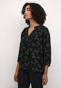 Blouse en tissu noir avec motif floral vert, décolleté en v et détails froncés au niveau du décolleté et des manches. Texture douce, coupe décontractée.