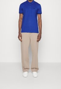 Královsky modré polo tričko s třemi knoflíky, černé logo na hrudi, spárováno s béžovými kalhotami širokého střihu a bílými teniskami. Hladká textura látky.