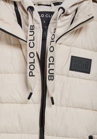 Beige polstret jakke med hætte, sort frontlynlås og snore med "POLO CLUB" tekst. Inkluderer et gummilogo-patch på brystet.