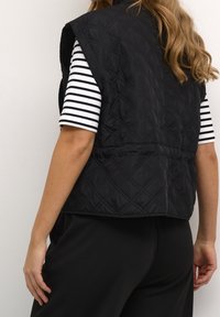 Schwarze gesteppte Weste mit hohem Kragen und ärmellosem Design, ausgestattet mit einer glatten Textur und horizontalen Nähten, über einem gestreiften Hemd getragen.