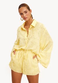 Mujer con una camisa amarilla de botones, suelta y texturizada, y pantalones cortos a juego con cordón, de pie con las manos en los bolsillos frente a un fondo blanco.