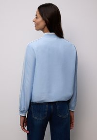 Mujer con cabello largo y oscuro, vistiendo una chaqueta bomber azul claro y jeans azul oscuro, de pie contra un fondo blanco liso, mirando de lado.