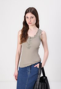 Geribbelde tanktop in licht olijfgroen met kantafwerking en een knoopdetail bij de halslijn, gecombineerd met een spijkerrok en een zwarte totebag.