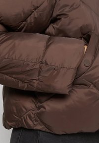 Doudoune marron avec un design matelassé, en tissu brillant, et un fermoir à pression près du poignet. Texture douce avec une finition légèrement brillante.