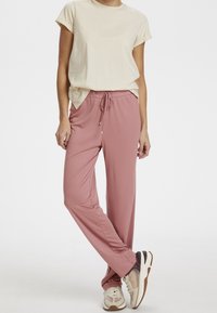 Svetle béžová polokošile s krátkým rukávem spárovaná s měkkými růžovými tepláky, které mají stahovací pás a volný střih. Celý outfit doplňují casual bílé tenisky.