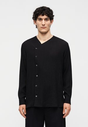 Νέος άνδρας με σγουρά μαλλιά που φοράει μαύρο μακρυμάνικο πουκάμισο με ασύμμετρο ντεκολτέ, στέκεται μπροστά από απλό φόντο.