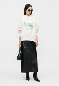 Γυναίκα που φοράει λευκό φούτερ με κουκούλα με πράσινο τύπωμα «Döner House», μαύρη δερμάτινη midi φούστα, μαύρα μπότες με τακούνι, γυαλιά ηλίου και κρατάει μια μικρή μαύρη τσάντα.