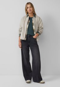 Giacca bomber grigia chiara sopra una maglietta verde a coste e una camicia a scacchi, abbinata a jeans a gamba larga scuri e sneakers crema. Design pulito e minimalista.