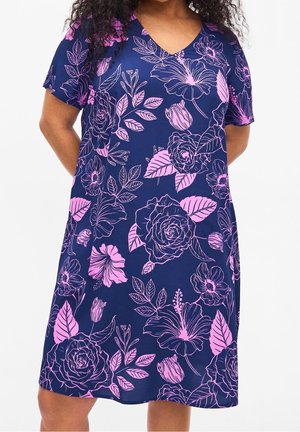Frau trägt ein marineblaues Kleid mit kurzen Ärmeln und großem rosa Blumenmuster, steht mit den Händen hinter dem Rücken.