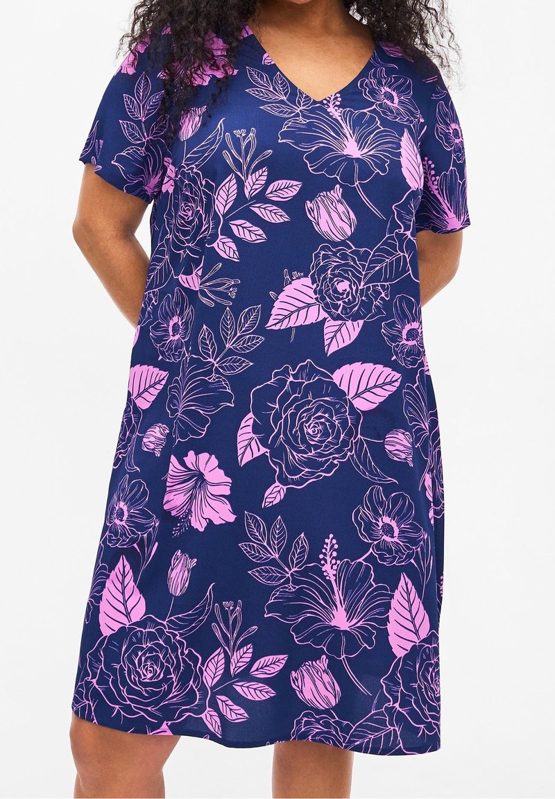 Femme portant une robe bleu marine à manches courtes avec un grand imprimé floral rose, debout, les mains derrière le dos.