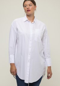 Camisa blanca de algodón con botones, con un bolsillo en el pecho, mangas largas y dobladillo redondeado. Textura suave con un diseño minimalista.