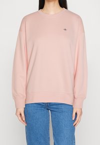 Sudadera rosa claro, cuello redondo, mangas largas con puños de canalé, corte relajado, pequeño logo en el pecho, combinada con jeans azules.