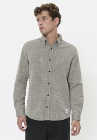 Cămașă cu mânecă lungă de culoare bej deschis, cu un model geometric negru, guler cu butoane și butoane negre. Include un etichetă pe marginea inferioară.