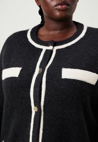 Μαύρο και κρεμ καρδιογκάλι, φτιαγμένο από μαλακό πλεκτό υλικό, με στρογγυλή λαιμόκοψη, μεγάλους κουμπιά και αντιθετικά υφασμένα τσέπες.