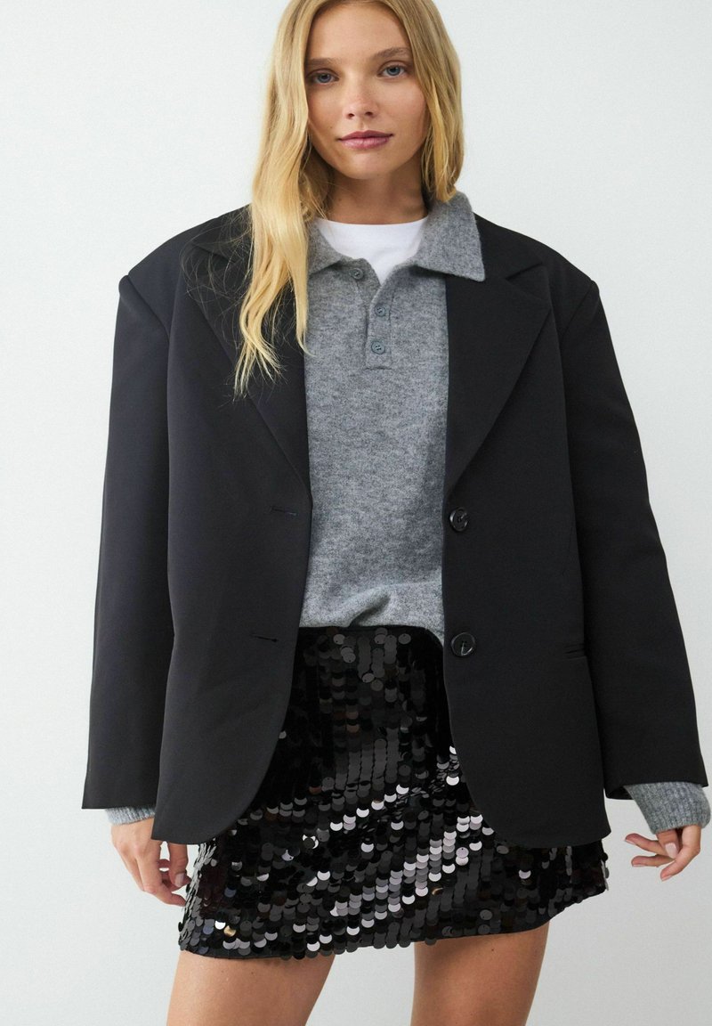 Czarny oversize blezer, szary sweter z kołnierzem oraz błyszcząca czarna spódnica mini pokryta cekinami. Blezer posiada dwie guziki i ma wyraźnie zarysowany krój.