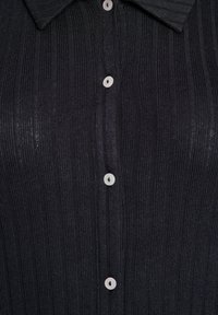 Černý žebrovaný pletený svetr s špičatým límcem a světlými knoflíky vpředu. Textura vypadá měkce a mírně leskle.