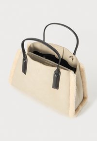 Beige håndtaske med bløde, pelslignende sider, sorte læderhåndtag og en lynlåslukket indre lomme. Glat tekstur og struktureret design.