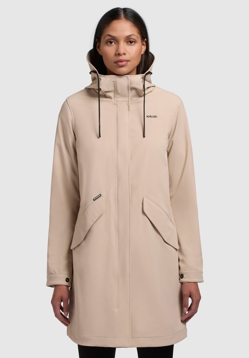 Beige vattentät jacka med hög krage, dragsko på huva, framsida med dragkedja och två stora fickor. Slät textur med en minimalistisk design.