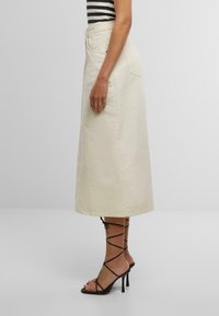 Una gonna di jeans color crema con vita alta, caratterizzata da una lunghezza completa e sottili dettagli di cucitura. Abbinata a sandali con tacco neri e con cinturini.