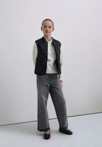 Musta, tikattu liivi valkoisen, ribbineuletun neuleen päällä, yhdistettynä väljien harmaiden farkkujen kanssa, joissa on koristeita ja halkiot lahkeissa. Mustat niititetyt ballerinat.