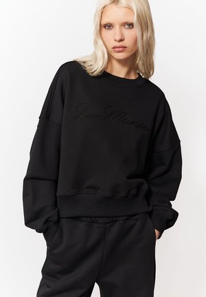 Szőke nő egyenes hajjal, fekete, túlméretezett pulóvert és hozzáillő fekete melegítőnadrágot visel, kezét a zsebében tartja.