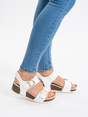 Legs in blue skinny jeans wearing white wedge sandals with gold buckles, standing on a white surface.