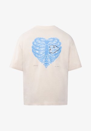 T-shirt beige avec un graphique de cage thoracique bleu en forme de cœur et un petit château blanc dans la zone de la poitrine, le texte indique « île des battements de cœur ».