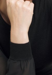 Svart tröja med ribbade muddar, med en slät yta och figursydd design. Hudfärgad hand knuten nära hakan.