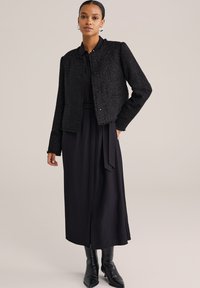 Naine, kes kannab tekstuurset musta lühikest jakki musta vöödiga midi kleidi peal, mustade teravate saabastega, seistes ühetoonilise tausta ees.