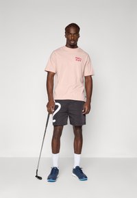 Lyserød t-shirt med rødt logo, sorte shorts med hvidt nummer 2, marineblå sko og hvide sokker, mens han/hun holder en golfkølle.