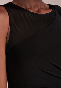 Svart ärmlös topp med en slät, strukturerad halsringning, genomskinligt material vid axeln och draperad tygdetalj på sidan.