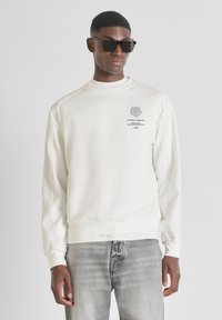 Sudadera blanca con un cuello alto, hombros caídos y puños acanalados. Presenta un estampado gráfico con texto en el pecho. Combinada con jeans grises.