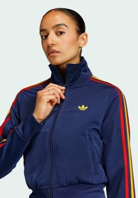 Sportska jakna tamnoplave boje s visokim ovratnikom, patentnim zatvaračem sprijeda, žutim logotipom, crvenim i žutim prugama duž rukava te elastičnim pojasom.