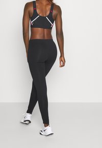 Svart sportbh med nättextur och röda detaljer, ihop med helsvarta leggings och vita sportskor, sedd bakifrån.